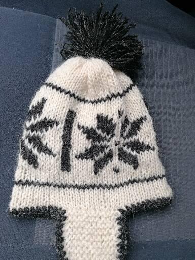 Ręcznie robiona, kremowa czapka z czarnymi, geometrycznymi wzorami i pomponem, leżąca na szarym tle.