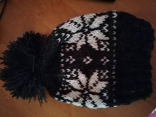 Czarno-biała czapka zimowa z pomponem i wzorem w śnieżynki, leżąca na drewnianym blacie.