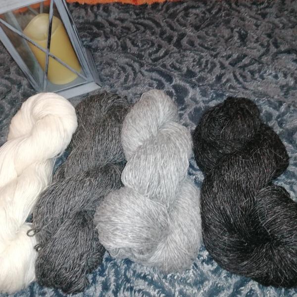 Cztery motki wełny w odcieniach bieli, szarości i czerni, ułożone obok siebie na wzorzystym, ciemnym tle, z lampionem ze świecą w tle.