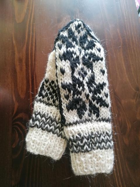 Para dzierganych, wełnianych mitenek w kolorze ecru i czarnym, z geometrycznym wzorem, leżących na drewnianym stole.