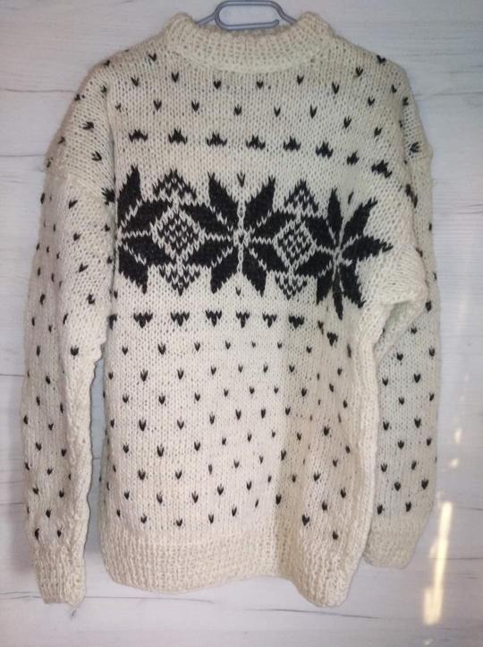 Ręcznie robiony, kremowy sweter z czarnymi wzorami w kształcie płatków śniegu i kropek, wiszący na plastikowym wieszaku na tle jasnego, drewnianego panelu.