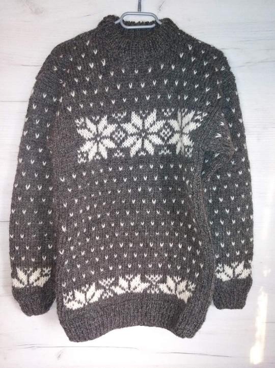 Szary, wełniany sweter z białymi wzorami śnieżynek, zawieszony na wieszaku na tle białej, drewnianej ściany.