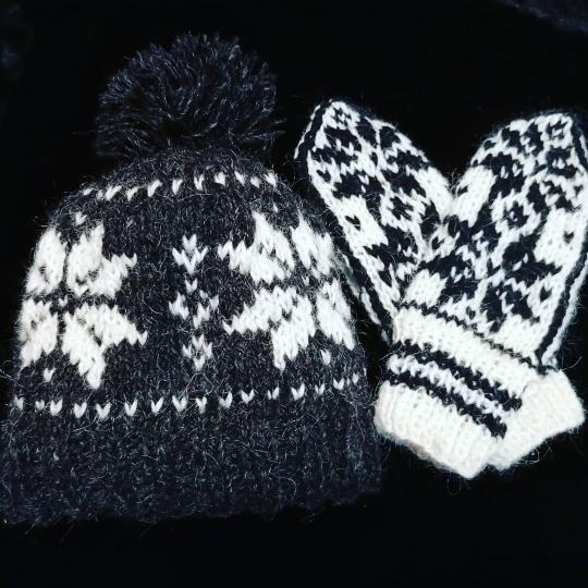 Ręcznie robiona czapka z pomponem i para dzierganych rękawic z wzorem w kolorach czerni i bieli, leżące na czarnym tle.