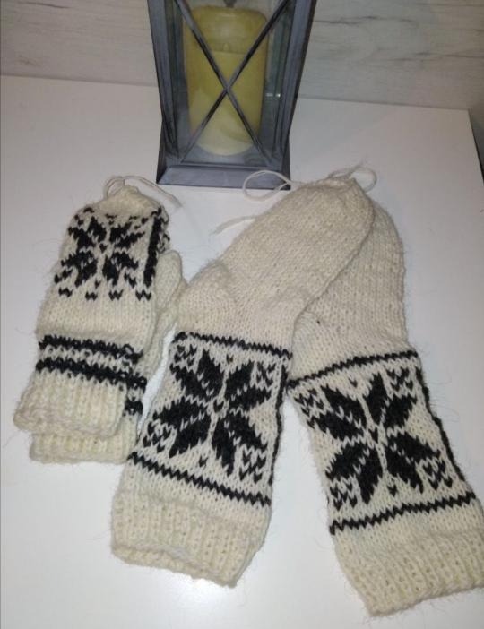 Ręcznie robiony komplet: wełniane skarpety i rękawiczki w kolorze ecru z czarnym wzorem śnieżynek, leżące obok metalowej latarenki ze świecą.