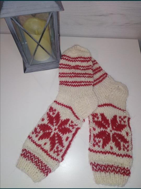 Para kremowych, ręcznie robionych wełnianych skarpet z czerwonymi wzorami śnieżynek i paskami, leżąca na białym stole obok szarej latarenki ze świecą w środku.