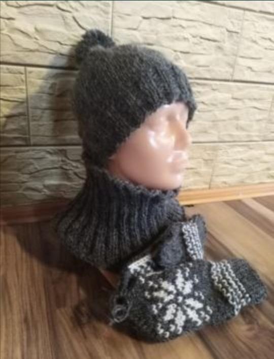 Szary, ręcznie robiony komplet zimowy: czapka z pomponem, komin i rękawiczki z motywem śnieżynki, prezentowany na manekinie na tle ściany z cegły i drewnianej podłogi.