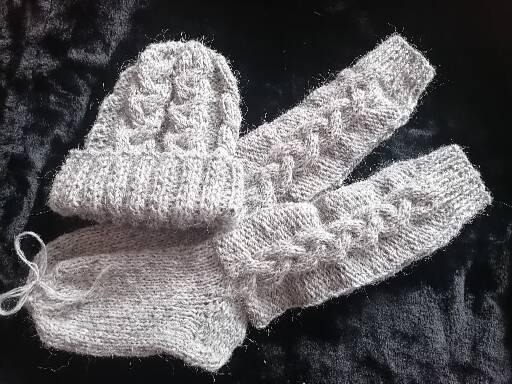 Ręcznie robiony komplet: szara czapka z warkoczowym wzorem i ocieplacze na ręce, leżące na ciemnym, teksturowanym tle.