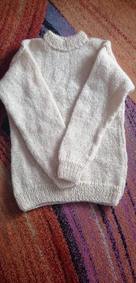 Ręcznie robiony, kremowy sweter z grubym splotem, leżący na kolorowej, wzorzystej tkaninie. Widoczne detale splotu i wykończenia.