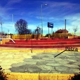 Ułożona nawierzchnia z szarej kostki brukowej w kształcie nieregularnych płyt, otoczona żółtą obwódką, prowadząca do schodów z czerwonej kostki. W tle widoczny budynek z reklamą.