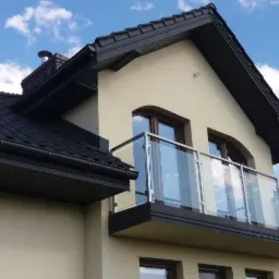 Elewacja domu z balkonem zabezpieczonym szklaną balustradą na tle błękitnego nieba z chmurami.