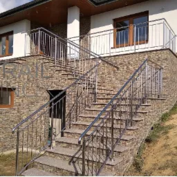 Schody zewnętrzne z balustradą ze stali nierdzewnej, prowadzące do domu z elewacją z kamienia dekoracyjnego.