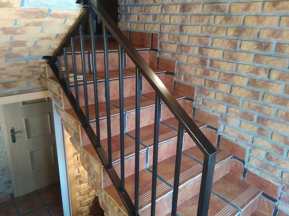 Schody wyłożone terakotą z ciemną, metalową poręczą na tle ceglanej ściany z widocznym wejściem do piwnicy.