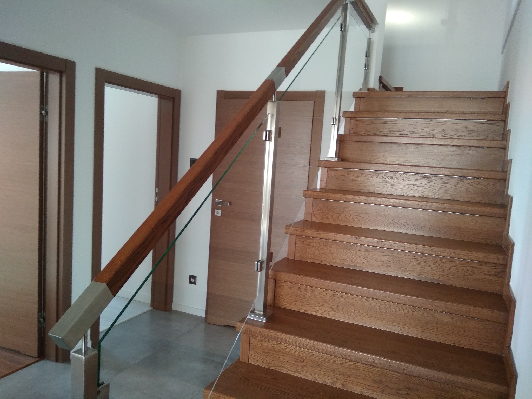 Szczegółowe ujęcie drewnianych schodów z nowoczesną balustradą: drewniana poręcz, szklane wypełnienie i stalowe słupki, widoczne drzwi w tle.