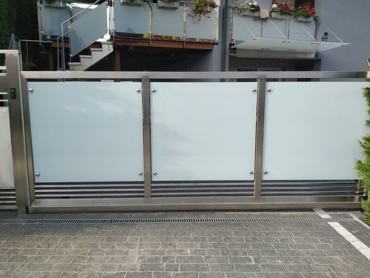 Przesuwna brama wjazdowa z mlecznego szkła i nierdzewnej stali, widoczna z zewnątrz na tle domu z balkonem i schodami z balustradą szklaną.