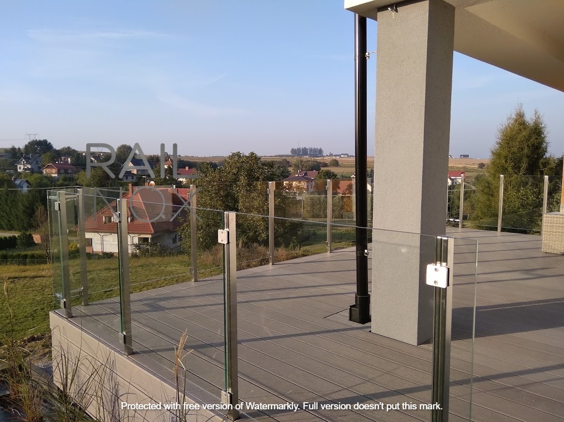 Szklana balustrada na tarasie z widokiem na wiejski krajobraz, słupki ze stali nierdzewnej, deski kompozytowe na podłodze.