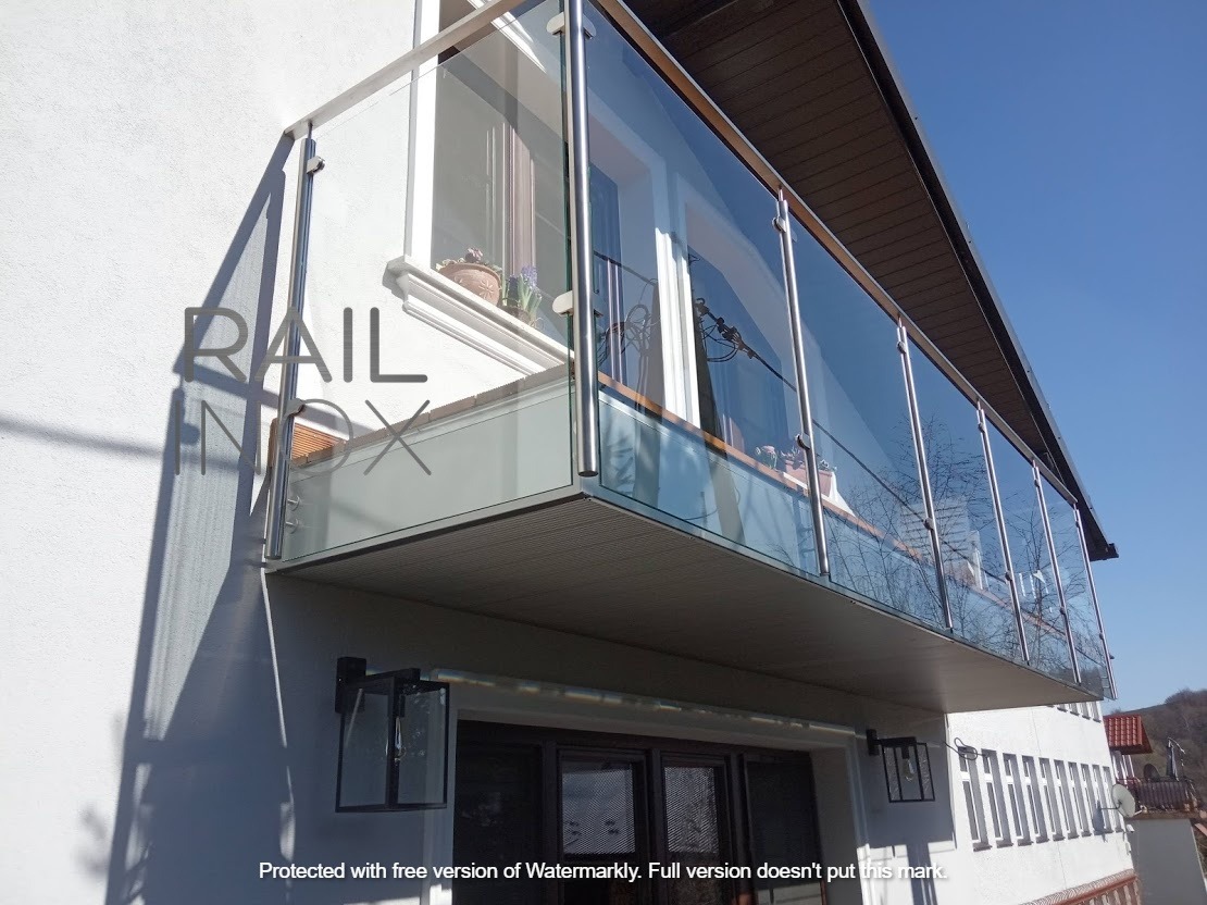 Szklana balustrada balkonowa z elementami ze stali nierdzewnej, widok z dołu na tle błękitnego nieba, z widocznymi lampami ściennymi w stylu retro.