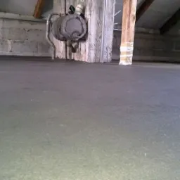 Świeżo wylana, gładka powierzchnia podłogi w surowym wnętrzu, widoczne fragmenty muru z pustaków, drewniany słup owinięty taśmą i instalacja grzewcza przy ścianie.