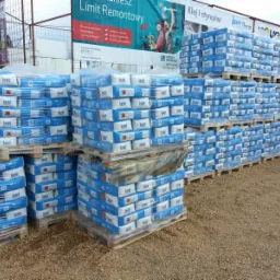 Palety z niebieskimi workami kleju, ustawione w stosy na placu budowy, reklama 'Klej i styropian' w tle.