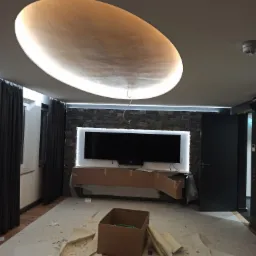 Owalna lampa sufitowa z podświetleniem LED, kamienna ściana z podświetlonym telewizorem, zabezpieczone meble kartonem, widok na remontowany pokój z drewnianą podłogą i zasłoniętym oknem.