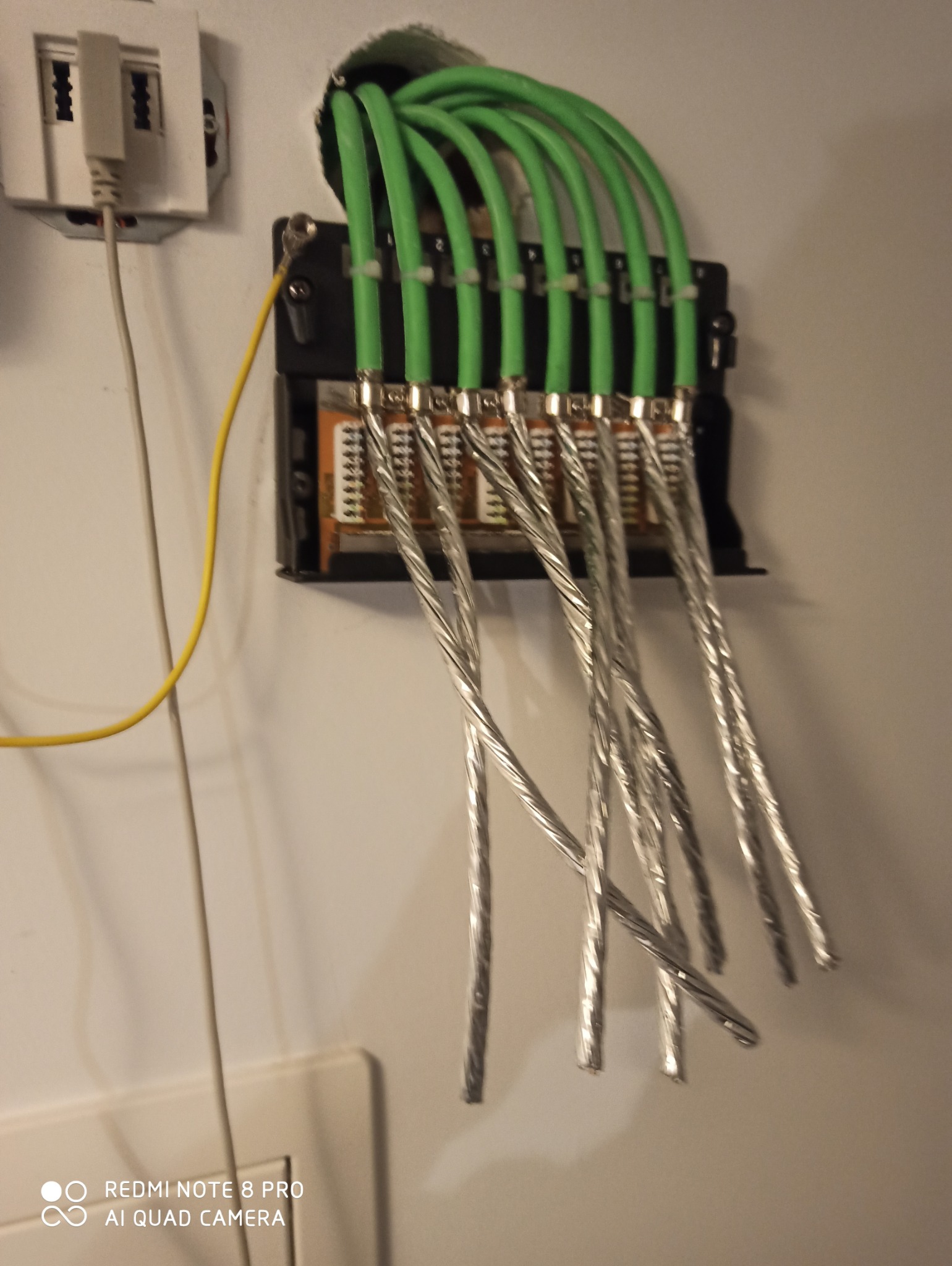 Zestaw podłączonych kabli sieciowych z zielonymi osłonami, zamocowanych do panelu na ścianie, z widocznym przewodem uziemiającym.