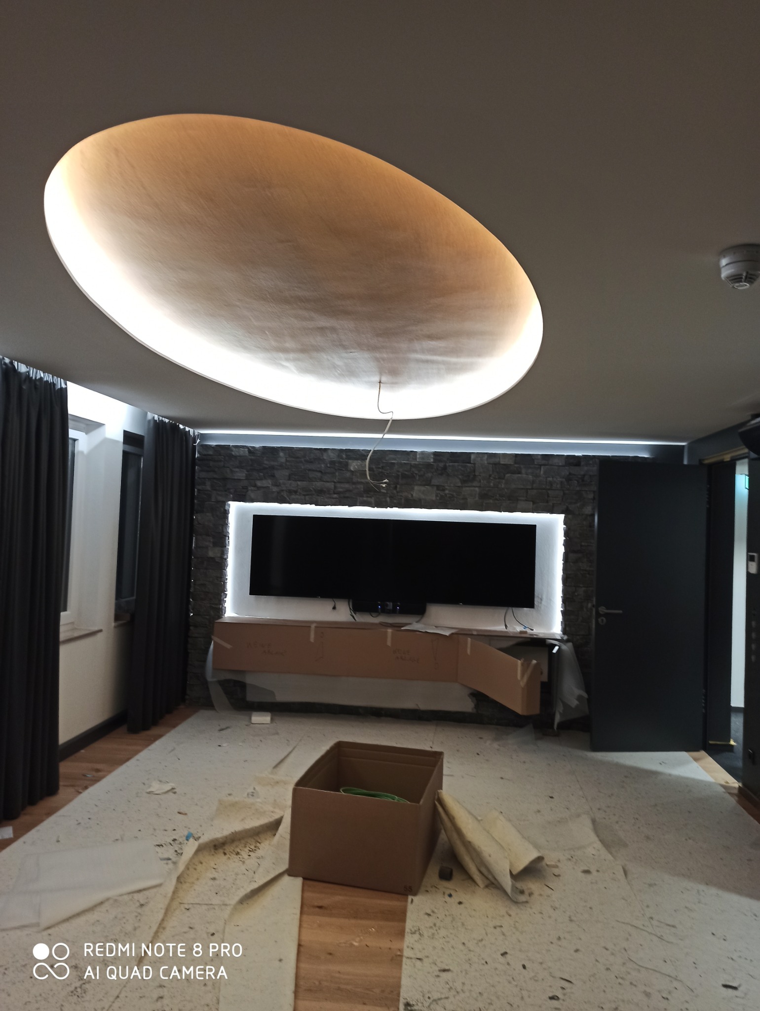 Owalna lampa sufitowa z podświetleniem LED, kamienna ściana z podświetlonym telewizorem, zabezpieczone meble kartonem, widok na remontowany pokój z drewnianą podłogą i zasłoniętym oknem.