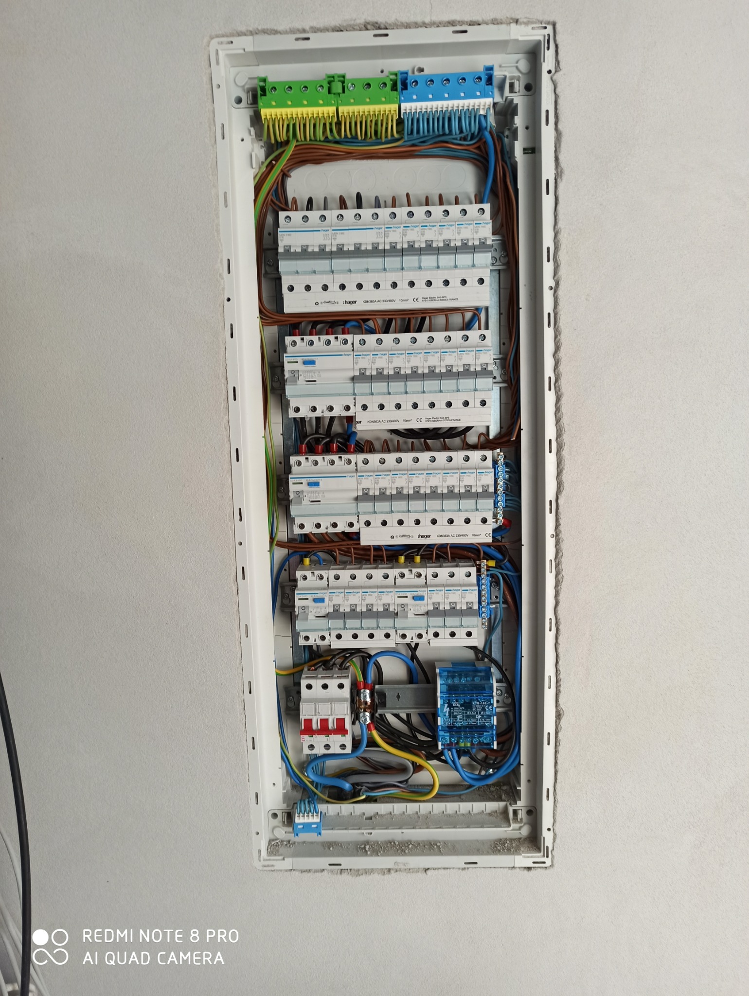 Rozdzielnica elektryczna Hager zamontowana w ścianie, widoczne bezpieczniki, kable w kolorowych izolacjach oraz listwy zaciskowe w odcieniach zieleni i niebieskiego. Ściana wokół rozdzielnicy...