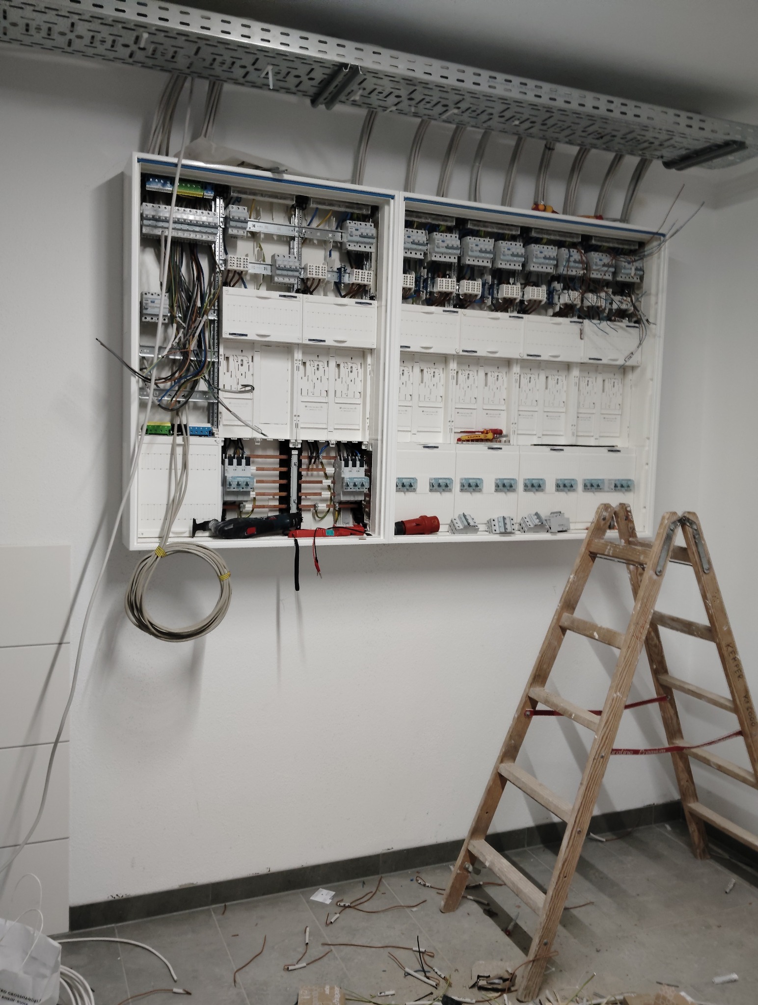 Rozbudowana skrzynka elektryczna w trakcie montażu z widocznymi przewodami, narzędziami i drabiną drewnianą na tle białej ściany w nowym budynku.