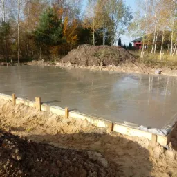 Świeżo wylany fundament pod budowę domu, z widocznym drewnianym szalunkiem i folią ochronną, na tle jesiennego krajobrazu z drzewami.