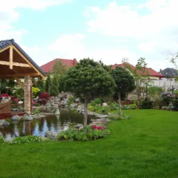 Starannie zaprojektowany ogród z oczkiem wodnym, kaskadą, drewnianą altaną i równo przystrzyżonym trawnikiem na tle zabudowy jednorodzinnej.