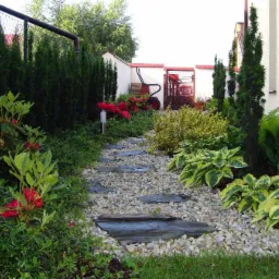 Ścieżka z kamiennych płyt prowadząca przez ogród z różnorodną roślinnością, w tle widoczna taczka i ogrodzenie.