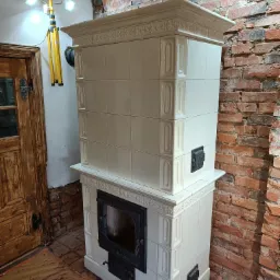 Biały, kaflowy piec z czarnymi drzwiczkami w surowym wnętrzu z czerwoną cegłą, oświetlony lampą budowlaną.