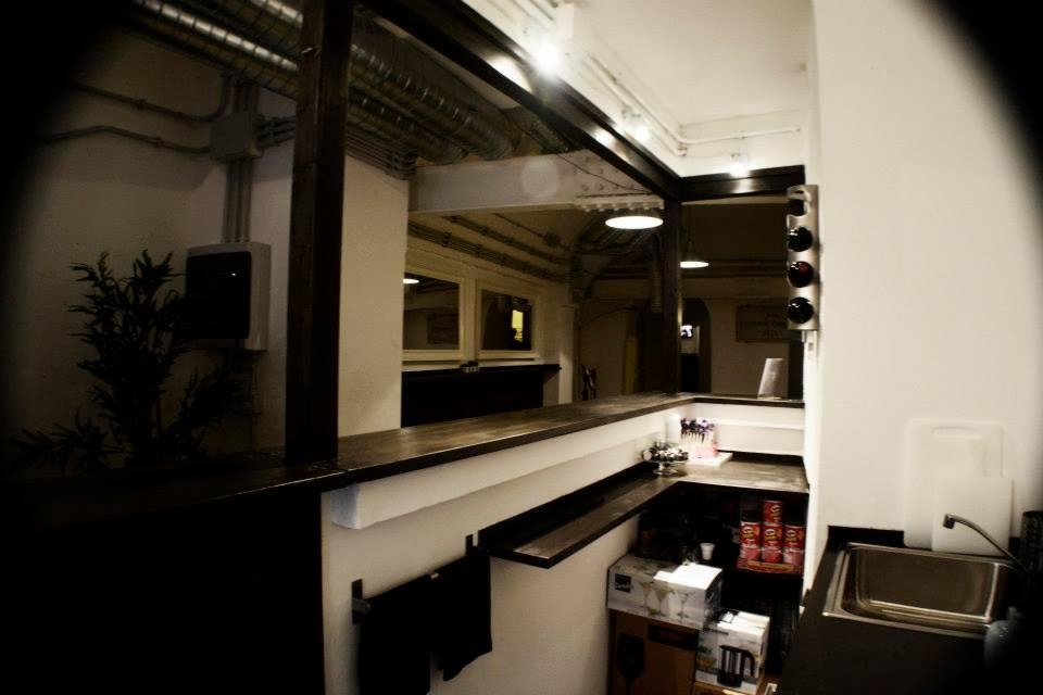 Loftowa aranżacja baru z blatem z ciemnego drewna, widokiem na dalszą część pomieszczenia i elementami instalacji na suficie. Minimalistyczny styl.