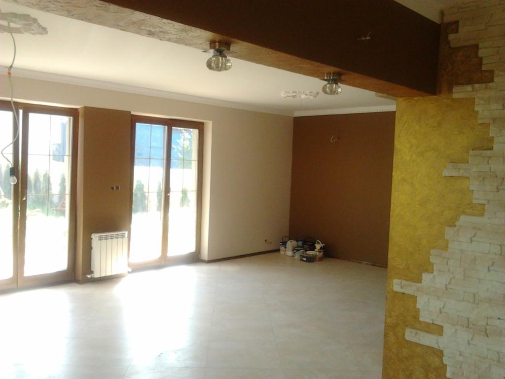 Wnętrze domu w trakcie wykańczania: ściana z dekoracyjnym tynkiem imitującym kamień, brązowa ściana pomalowana farbą, widoczne pojemniki z farbami, okna balkonowe z widokiem na ogród.