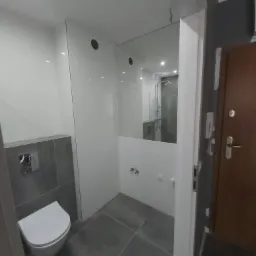 Nowoczesna łazienka po remoncie z wiszącą toaletą, szarymi płytkami podłogowymi i częściowo przeszkloną kabiną prysznicową, widoczna przez białą futrynę drzwi wejściowych do mieszkania.