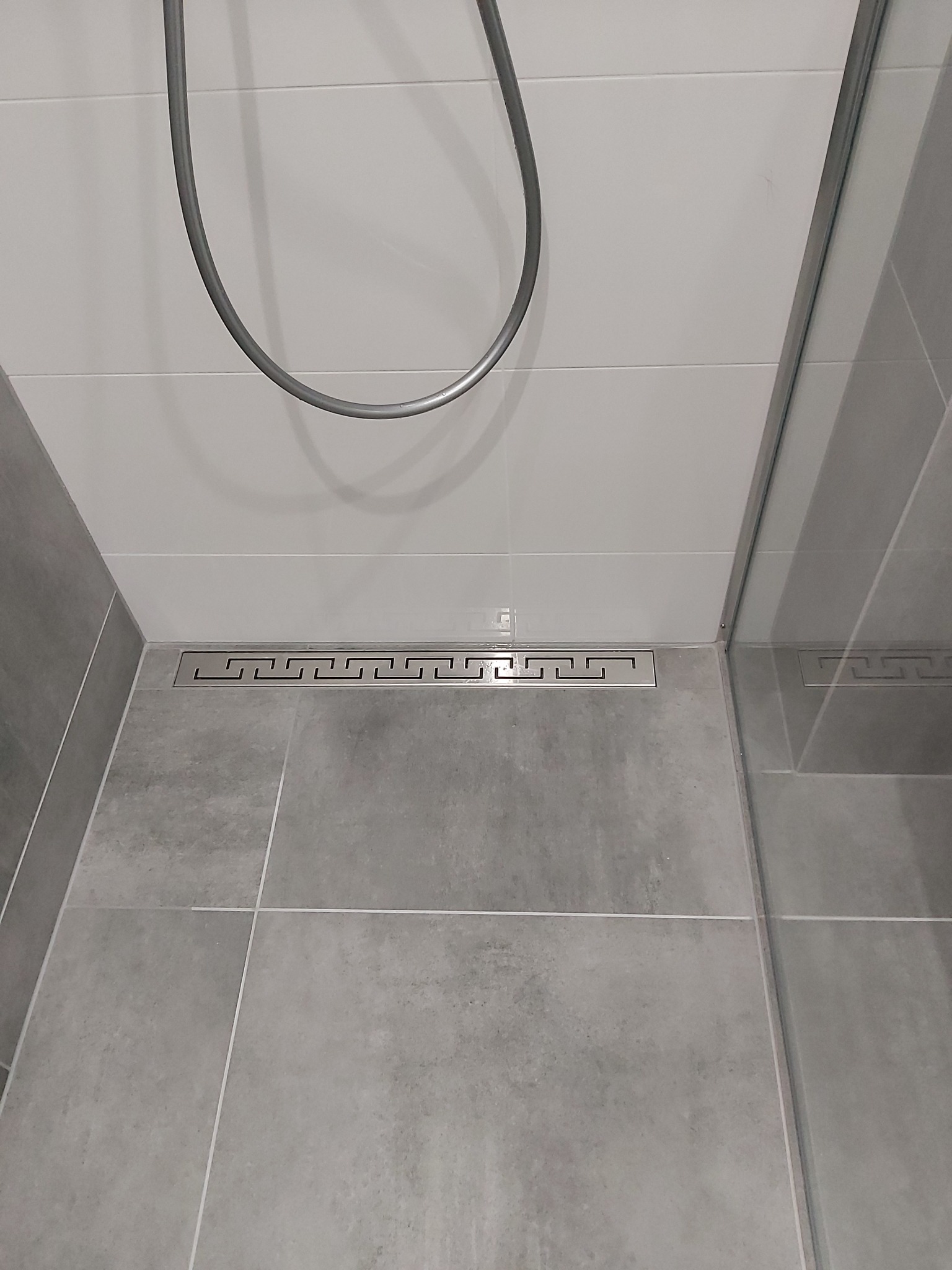 Szary prysznic z odpływem liniowym ozdobionym geometrycznym wzorem greckim, widoczna słuchawka prysznicowa i szklana kabina.
