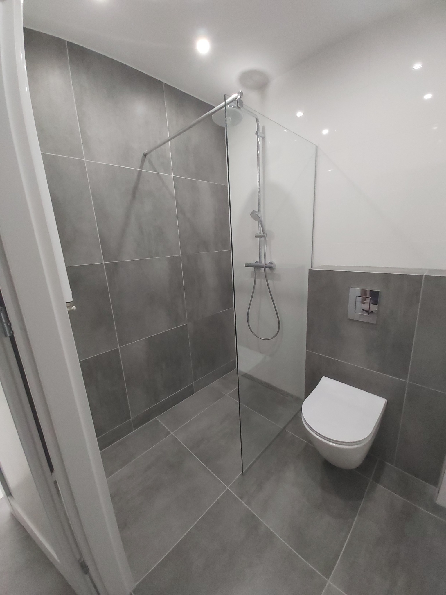 Nowoczesna łazienka z szarymi płytkami wielkoformatowymi, kabiną prysznicową typu walk-in i podwieszaną toaletą z chromowanym przyciskiem spłukiwania.