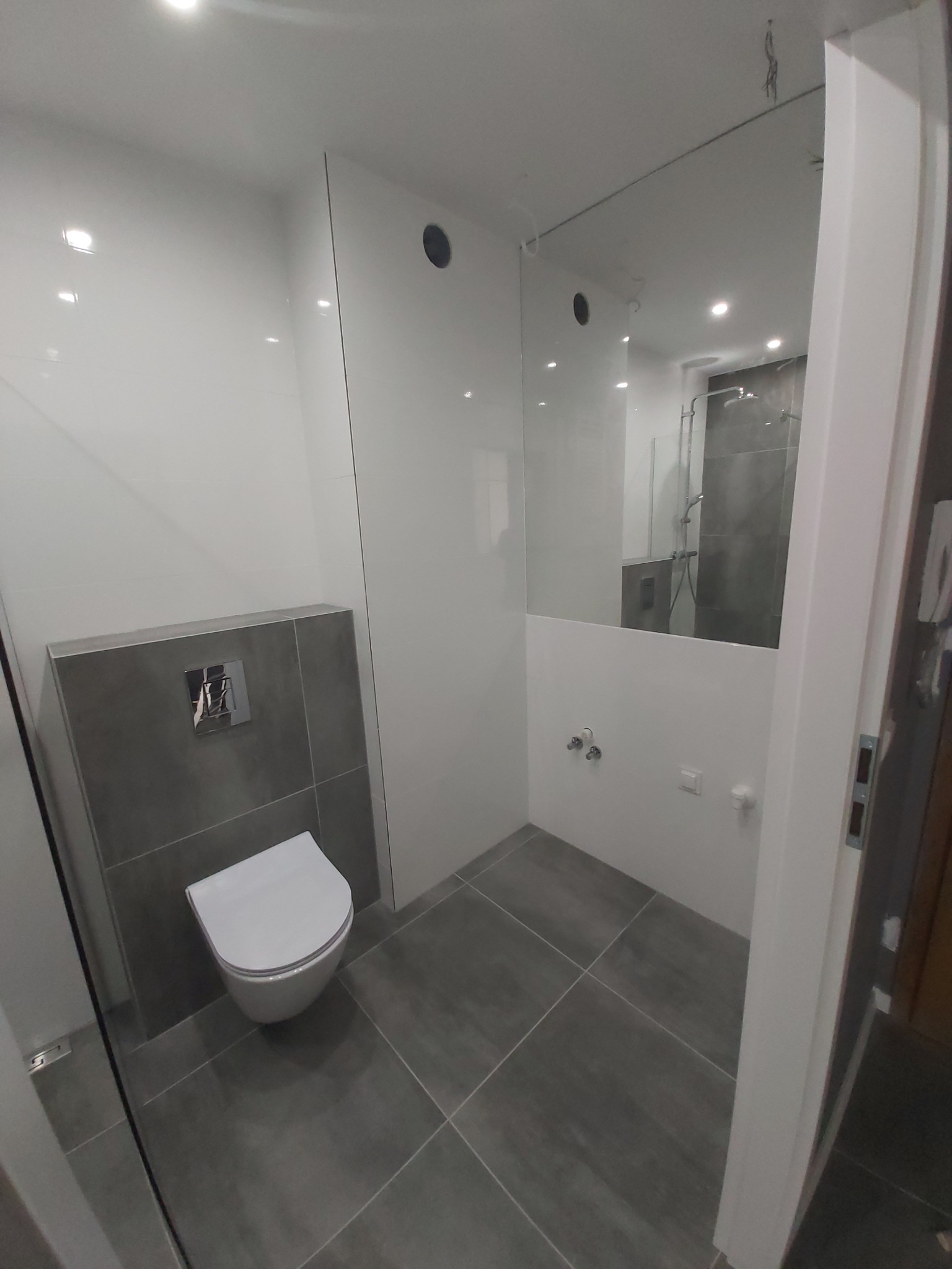 Nowoczesna łazienka z podwieszaną toaletą, szarą glazurą i lustrem powiększającym przestrzeń, w odbiciu widoczny prysznic.