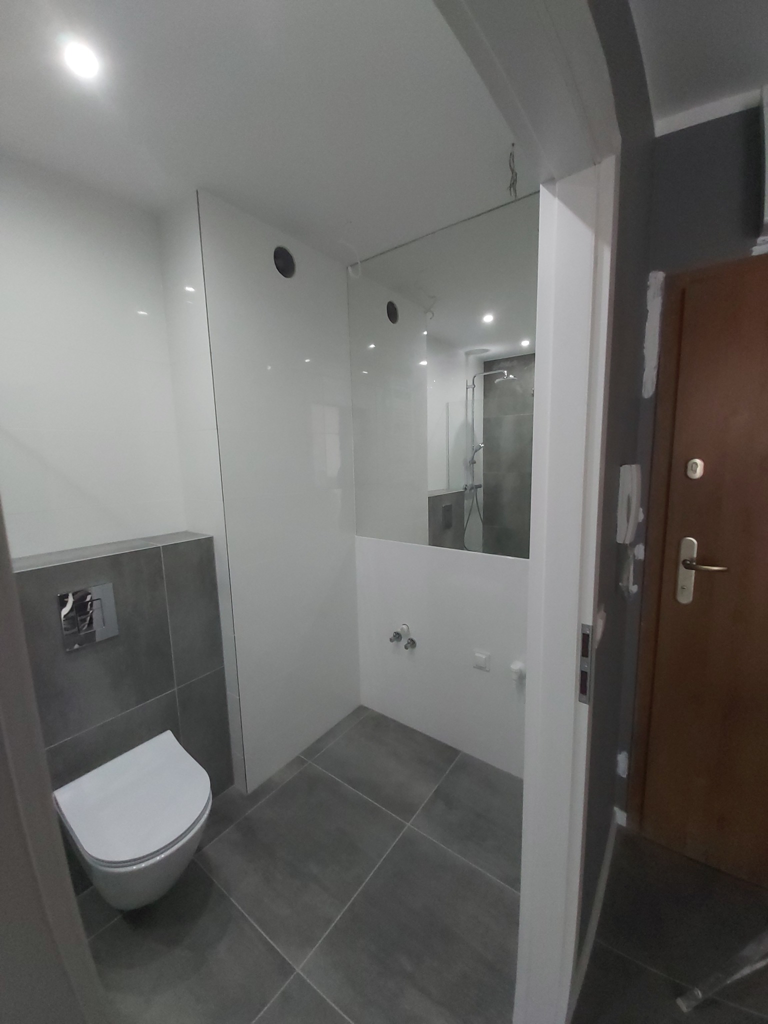 Nowoczesna łazienka po remoncie z wiszącą toaletą, szarymi płytkami podłogowymi i częściowo przeszkloną kabiną prysznicową, widoczna przez białą futrynę drzwi wejściowych do mieszkania.