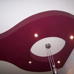 Dekoracyjny sufit podwieszany w burgundowym kolorze z okrągłym otworem i oświetleniem punktowym. W centrum wisi nowoczesna lampa.