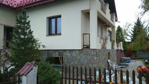 Dwupiętrowy dom z elewacją z jasnego tynku i kamienną podmurówką, otoczony zielenią i drewnianym ogrodzeniem. Widoczny fragment dachu z czerwonej dachówki.