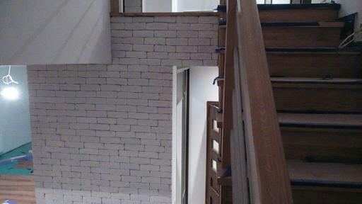 Klatka schodowa w trakcie wykończenia w Warszawie. Ściana z białej cegły, drewniane schody z widocznymi niedoskonałościami, surowy stan wnętrza.