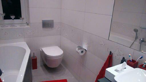 Nowoczesna, jasna łazienka z białymi płytkami zdobionymi motywem kwiatowym. Widoczna wanna, toaleta podwieszana i umywalka z lustrem.