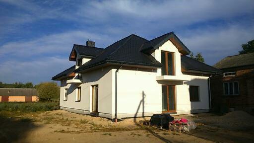 Nowoczesny, biały dom z czarnym dachem i drewnianymi oknami w słoneczny dzień. Elementy wykończeniowe czekają na dokończenie budowy. Widoczne materiały budowlane.