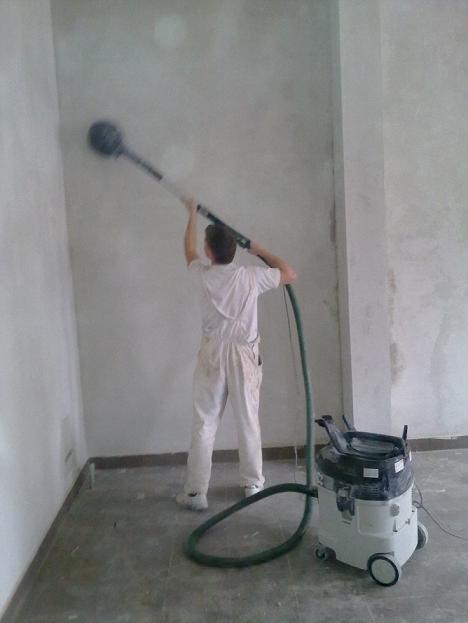 Pracownik w białym kombinezonie szlifuje ścianę szlifierką z odkurzaczem przemysłowym w rogu pomieszczenia, przygotowując ją do malowania.