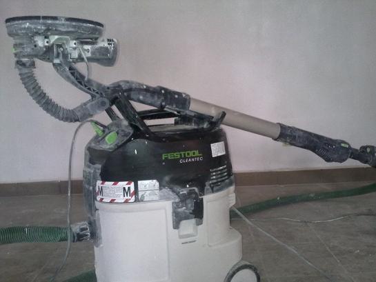 Szlifierka do gładzi marki Festool Cleantec z odkurzaczem, używana podczas prac wykończeniowych, pokryta pyłem gipsowym, z widocznym wężem odsysającym.