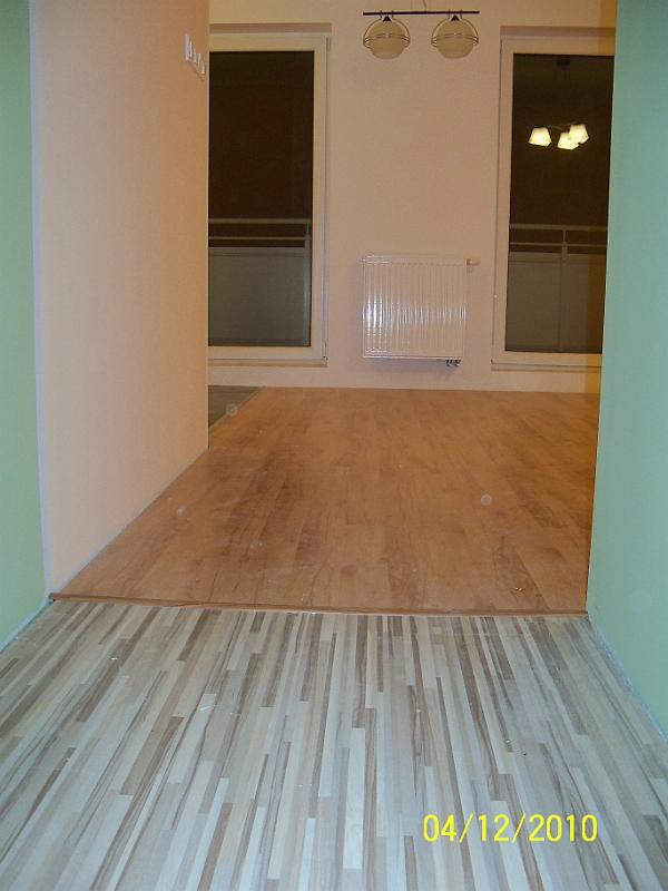 Układanie podłogi z paneli w Bydgoszczy, widoczny fragment nowej podłogi przechodzący w starszą, drewnianą podłogę w dalszej części pomieszczenia, ściany w odcieniach beżu i zieleni.