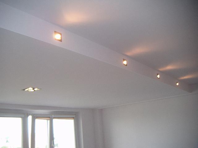 Biały sufit z zamontowanymi w listwie halogenowymi punktami świetlnymi, widoczne okno w tle.