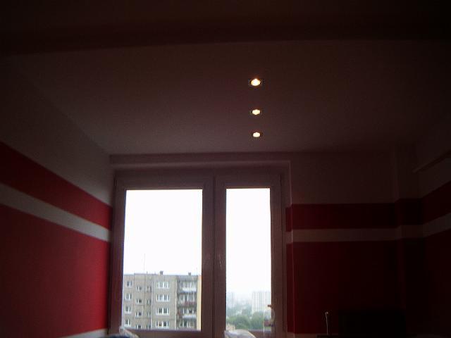Pokój po remoncie: Ściany pomalowane w poziome pasy czerwieni i bieli, sufit z trzema punktowymi lampami LED, okno z widokiem na miasto.