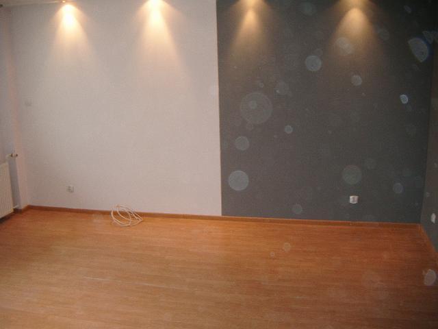 Pokój po remoncie z nową, drewnianą podłogą w odcieniu jasnego brązu, białą ścianą po lewej stronie i szarą ścianą po prawej oświetloną trzema punktowymi lampami sufitowymi, na podłodze leżą...