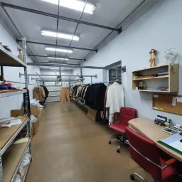 Wnętrze pracowni krawieckiej z rzędami ubrań na wieszakach, biurkiem z czerwonymi krzesłami i metalowymi regałami, oświetlone lampami jarzeniowymi.